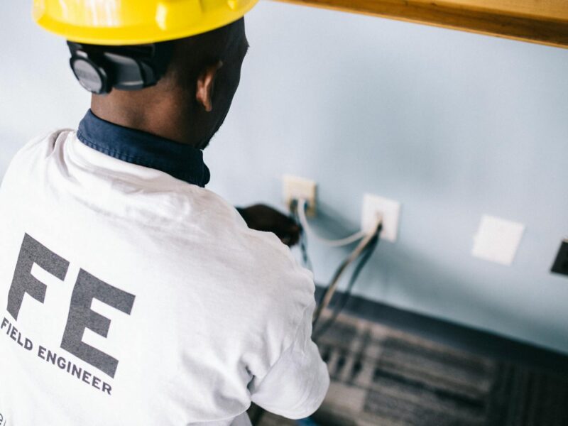 From above back view unrecognizable black field engineer wearing white shirt and protective hardhat sitting on floor and checking voltage in wall plugs