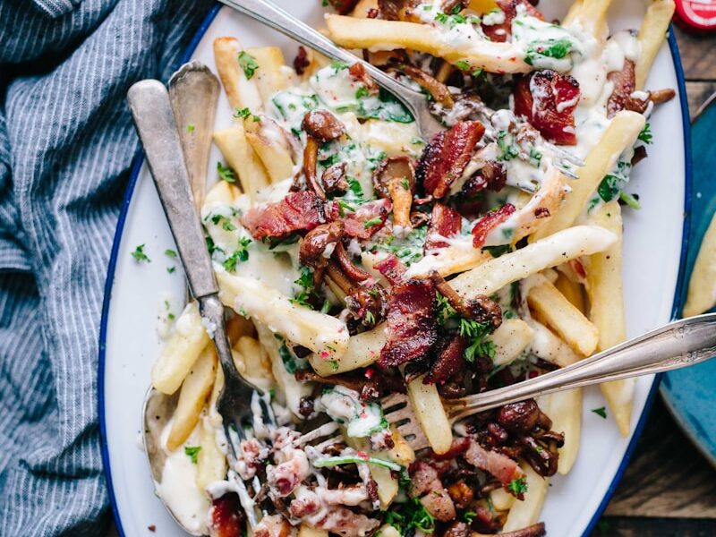 bacon strips and melted cheese topped fries on oval white and blue platter with gray stainless steel forks