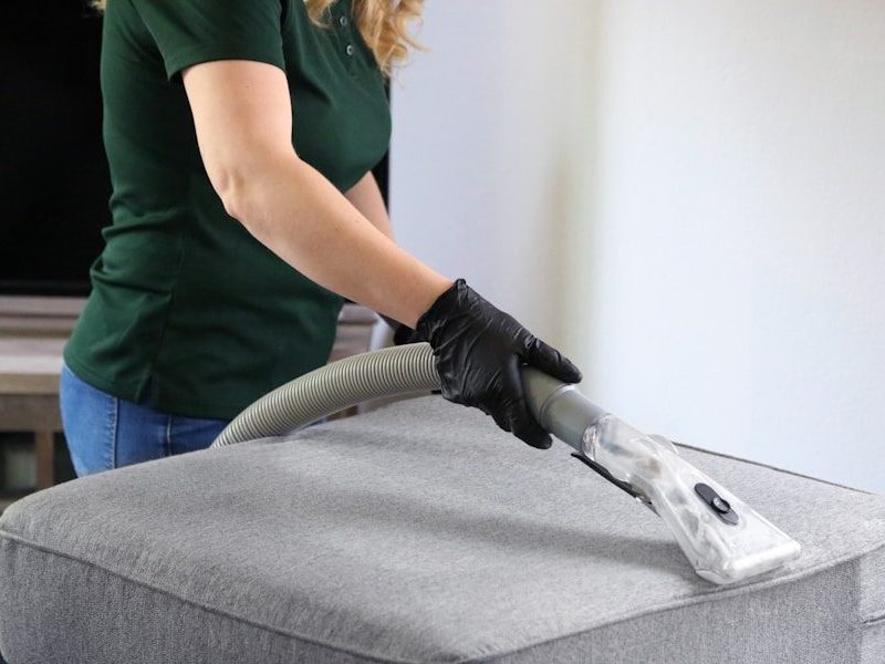a woman in a green shirt and black gloves vacuuming a gray ottoman