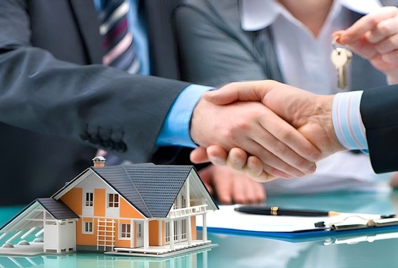 Two men shaking hands over a house model and keys.