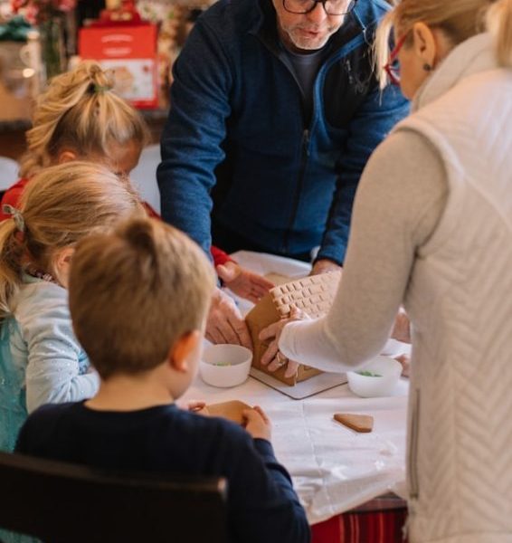 two women standing and three children sitting while preparing gingerbread house