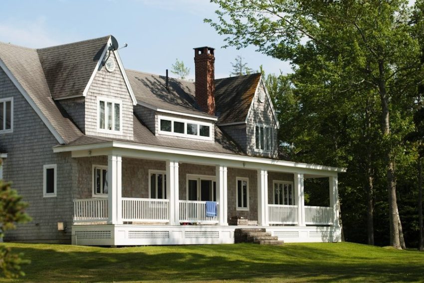A gray older two-story house with a stone chimney and a covered front porch set on a grassy lawn