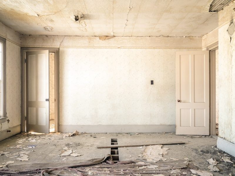 An abandoned room with peeling wallpaper, two open doors, and a damaged wooden floor