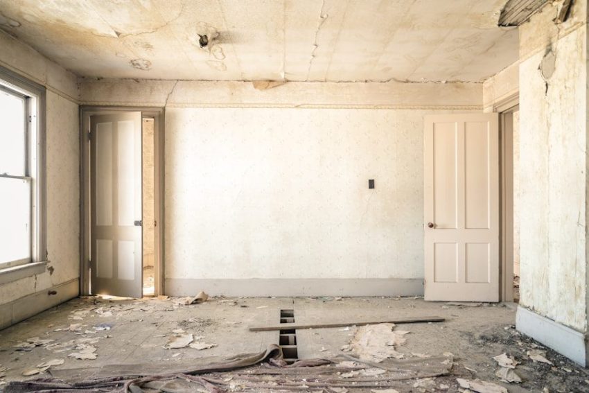 An abandoned room with peeling wallpaper, two open doors, and a damaged wooden floor
