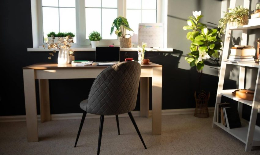 Home workspace with a wooden desk, quilted gray chair, laptop, lamp and plants beside a sunlit window.