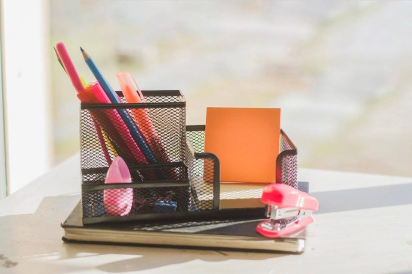 Black mesh organizer holding pens, pencils and orange notes on notebooks beside a pink stapler on a white desk.